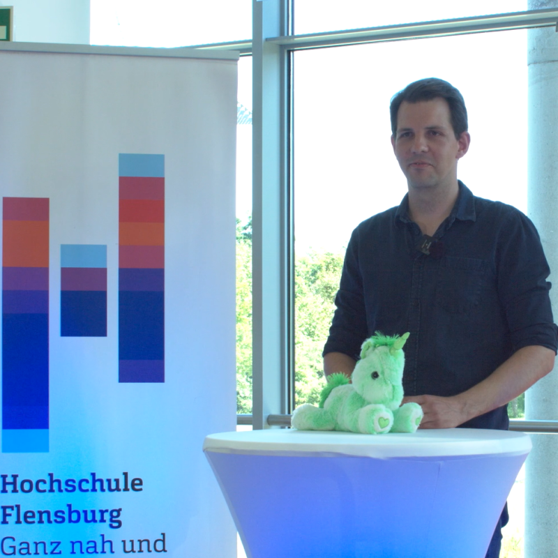 Ein junger Mann im schwarzen Hemd mit hochgekrempelten Ärmeln steht an einem Stehtisch vor einem Roll-Up mit dem Logo der Hochschule Flensburg und lächtelt in die Kamera.