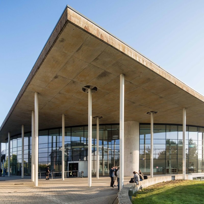 Das audimax-Gebäude auf dem Flensburger Campus: Eine verglaste Front, weiße Säulen und ein dreieckiges Dach, darüber blauer Himmel.