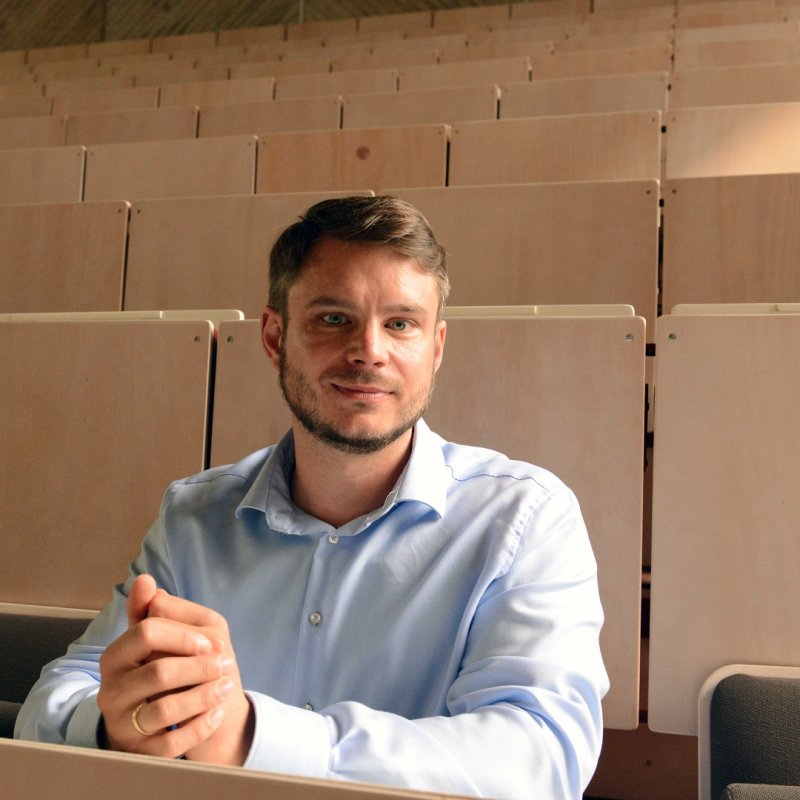 Ein junger Mann im weißen Hemd sitzt in einem leeren Hörsaal und blickt lächelnd in die Kamera.