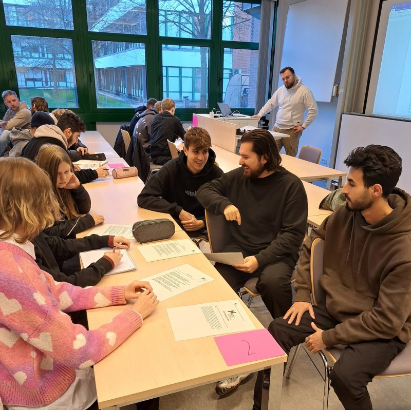 In einem Seminarraum sitzen sich mehrere Student*innen und Schüler*innen gegenüber und unterhalten sich, auf dem Tisch zwischen ihnen liegen Zettel und Stifte.