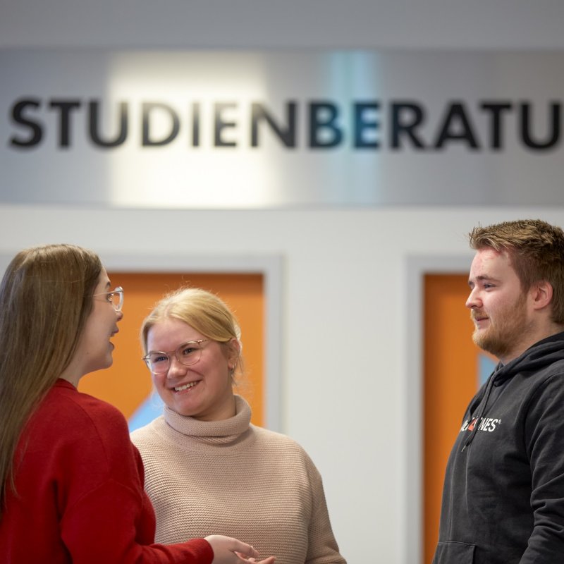Drei Studierende unterhalten sich lachend. Im Hintergrund sieht man zwei orangene Türen, über denen in metallischen Großbuchstaben das Wort "Studienberatung" steht.