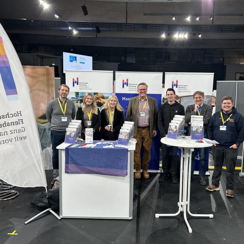 Sieben Personen verschiedenen Alters stehen hinter einem Messestand und blicken lächelnd in die Kamera. Hinter ihnen stehen Roll-Ups mit dem Logo der Hochschule Flensburg, vor ihnen eine Beachflag mit dem Logo. Auf Tischen vor ihnen stehen Aufsteller mit verschiedenen Flyern.