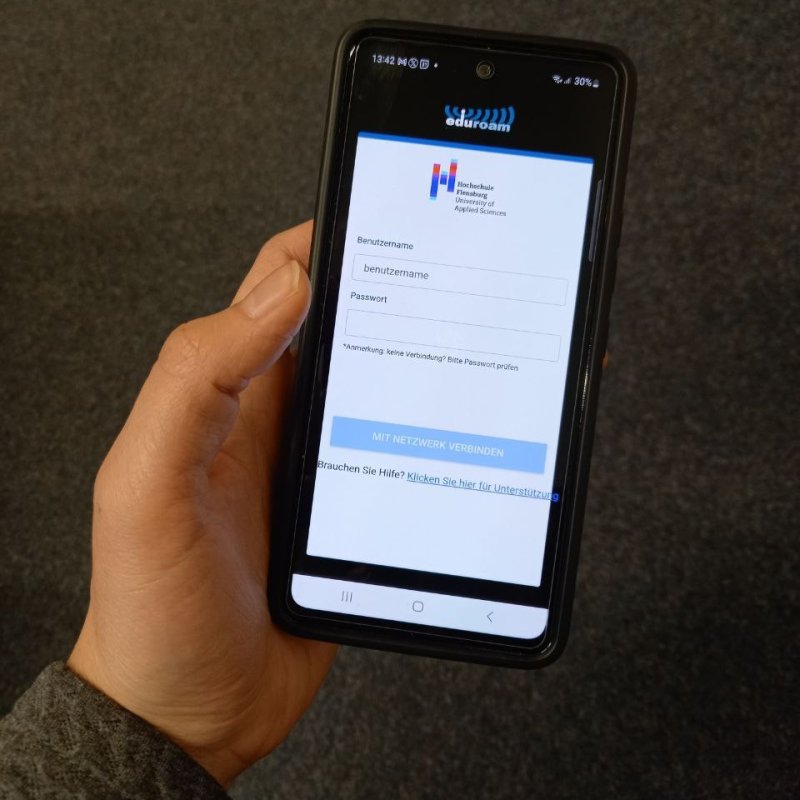 Eine Hand hält ein Smartphone, auf dem die Oberfläche einer App zu sehen ist. Oben steht "eduroam", darunter das Logo der Hochschule Flensburg, eine Eingabemaske und ein Button mit der Aufschrift "Mit Netzwerk verbinden".