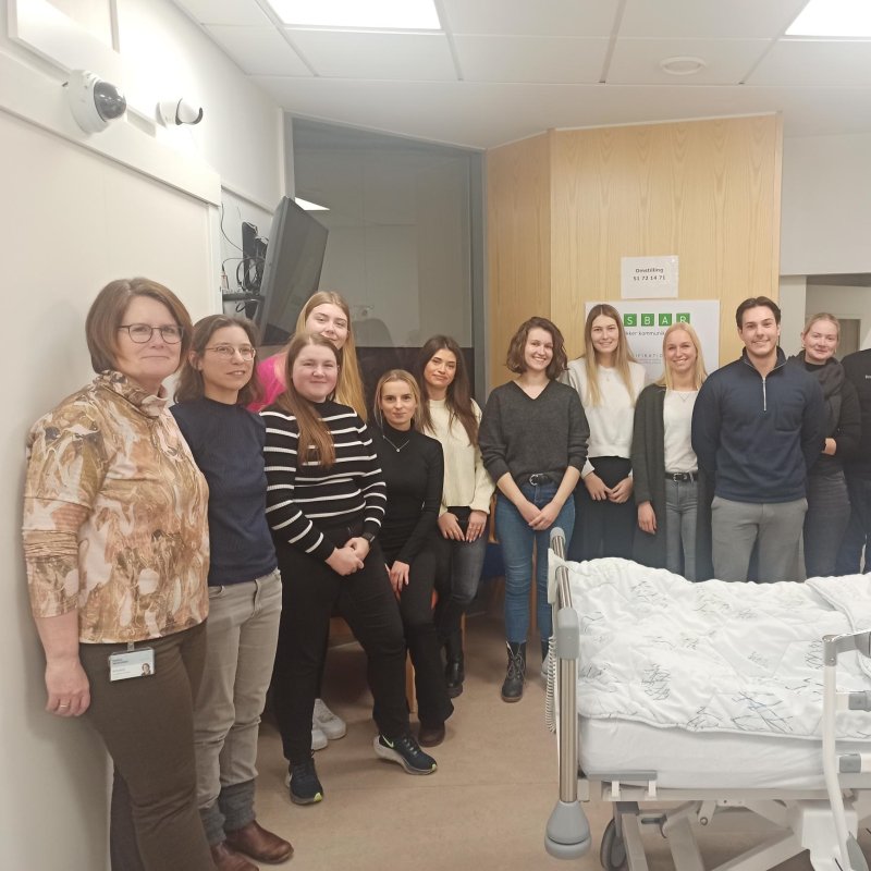 Eine Gruppe von jungen Menschen steht rund um ein Krankenhausbett, in dem eine Modellpuppe liegt. An der Wand hängt ein Monitor, neben dem Bett ein Tropf und an der Wand hinter den Studierenden hängt ein Poster mit Aufschrift in dänischer Sprache.