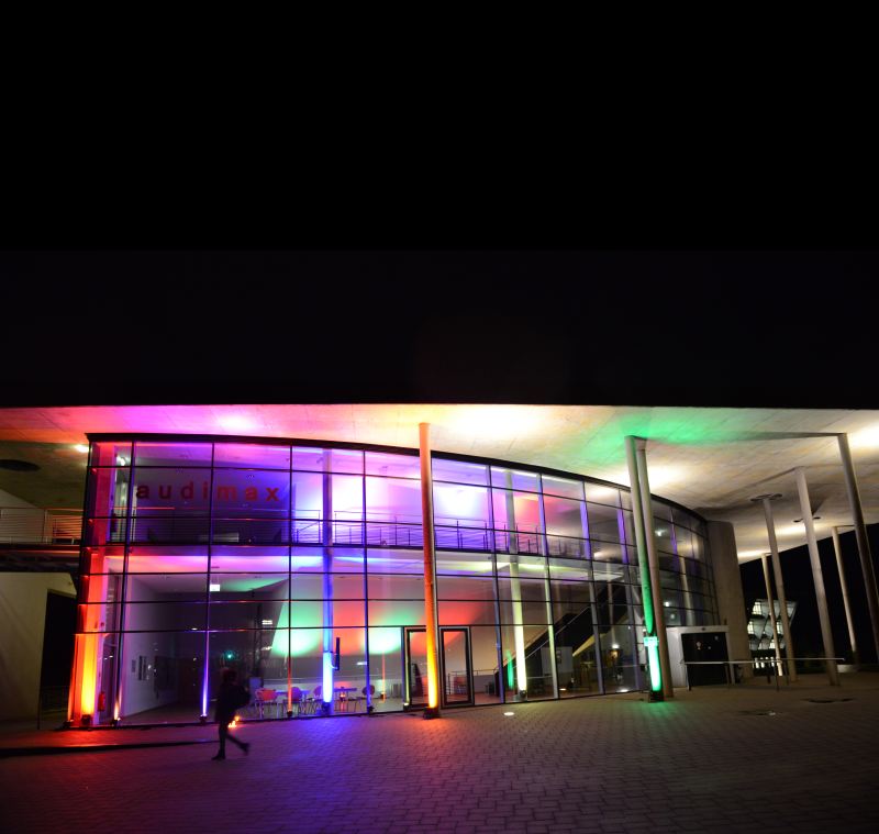 Das audimax der Hochschule Flensburg, ein moderner Bau mit Glasfront und dreieckigem Dach, wird im Dunklen in verschiedenen bunten Farben angeleuchtet.