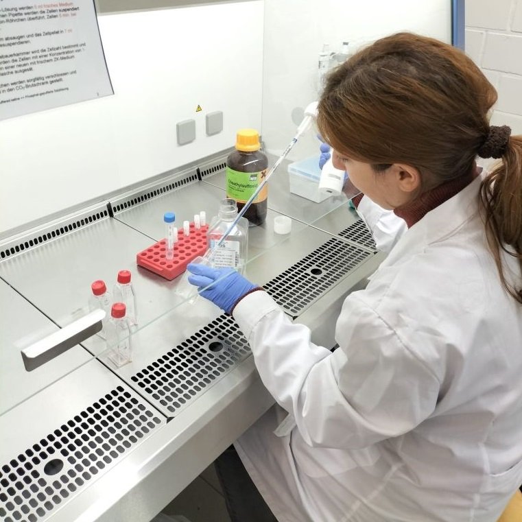 Eine junge Frau im Laborkittel sitzt an einem Labortisch und füllt mit einer Pipette eine Flüssigkeit in ein Glas ab.