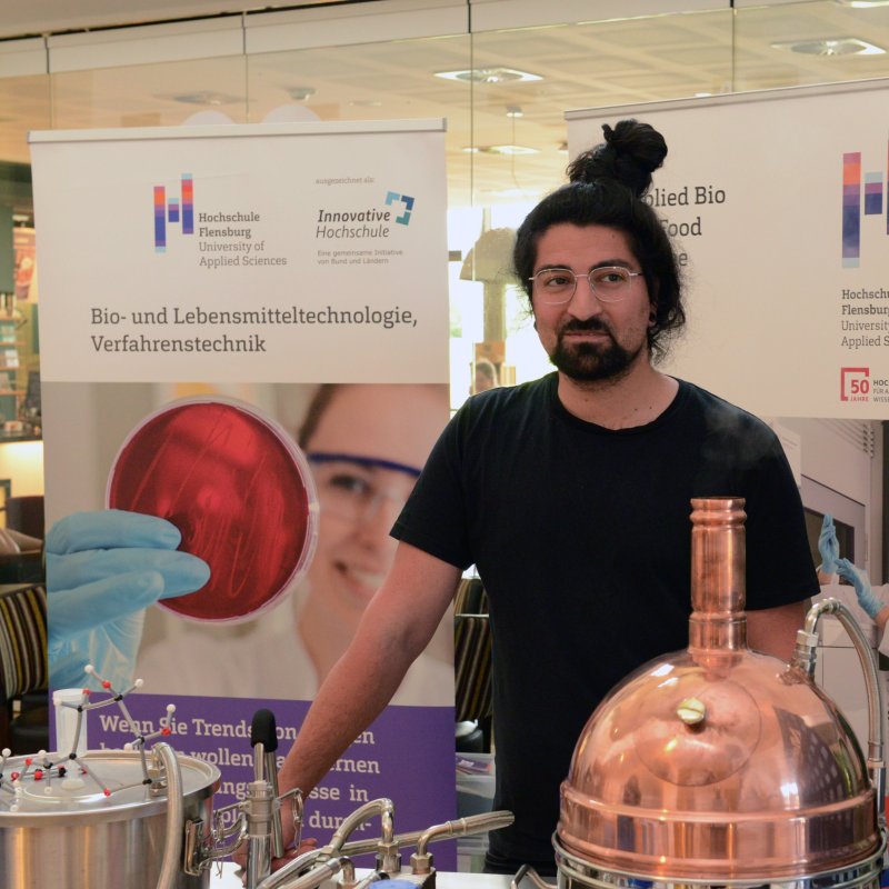 Ein Mann mit Bart und langen, zu einem Dutt gebundenen Haaren steht an einem Tisch, auf dem eine Versuchsapparatur mit einem silbenern und einem Kupferkessel aufgebaut ist. Im Hintergrund stehen zwei Roll-Ups mit dem Logo der Hochschule Flensburg, Fotos einer jungen Frau im Labor und der Aufschrift "Bio- und Lebensmitteltechnik, Verfahrenstechnik"