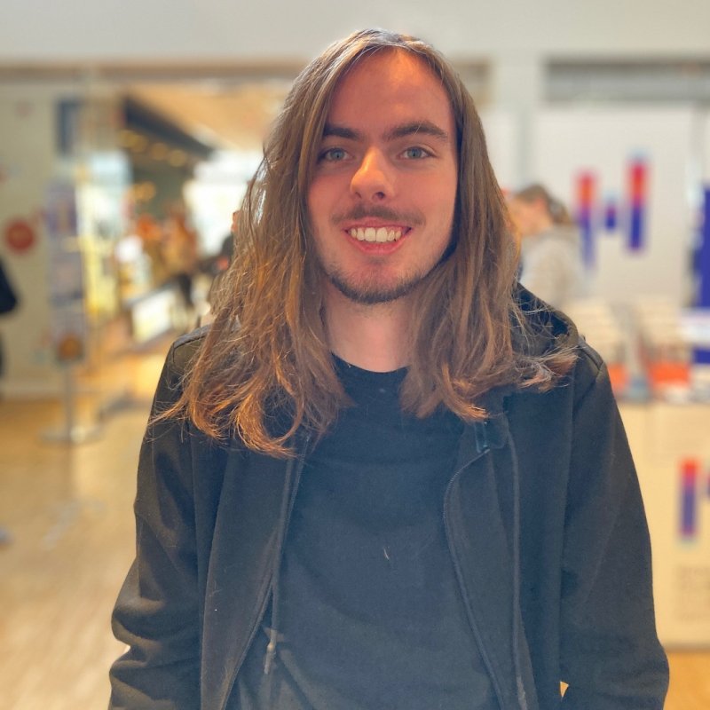 Ein junger Mann mit langen braunen Haaren, schwarzem T-Shirt und schwarzem Kapuzenpulli lächelt offen in die Kamera. Im Hintergrund sieht man verschwommen einen Infostand mit Flyern und dem Logo der Hochschule Flensburg.