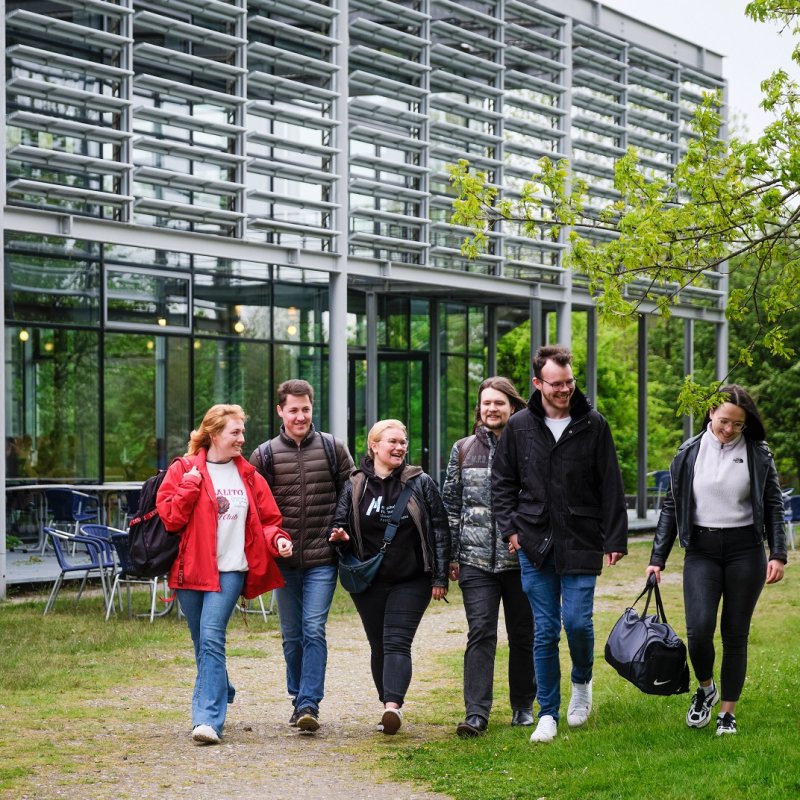 Eine Gruppe Studierender läuft einen Schotterweg auf einer grünen Wiese entlang. Im Hintergrund sieht man ein modernes Gebäude mit Glasfassade und einer Terrasse mit blauen Stühlen und Tischen: die Mensa des Flensburger Campus.
