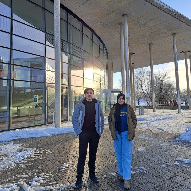 Ein junger Mann und eine junge Frau stehen vor dem Flensburger audimax: Ein modernes Gebäude mit geschwungener Glasfront, in der sich die Wintersonne spiegelt.