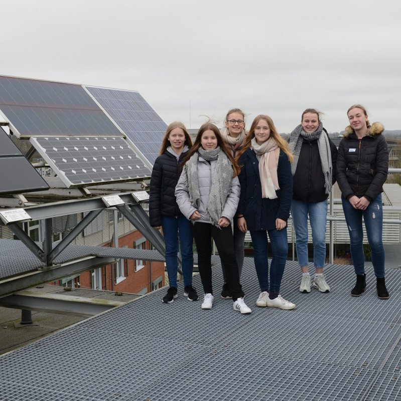 Eine Gruppe von Mädchen steht auf einem Flachdach neben einer Photovoltaik-Anlage, im Hintergrund sieht man in der Ferne Wiesen und Büsche.