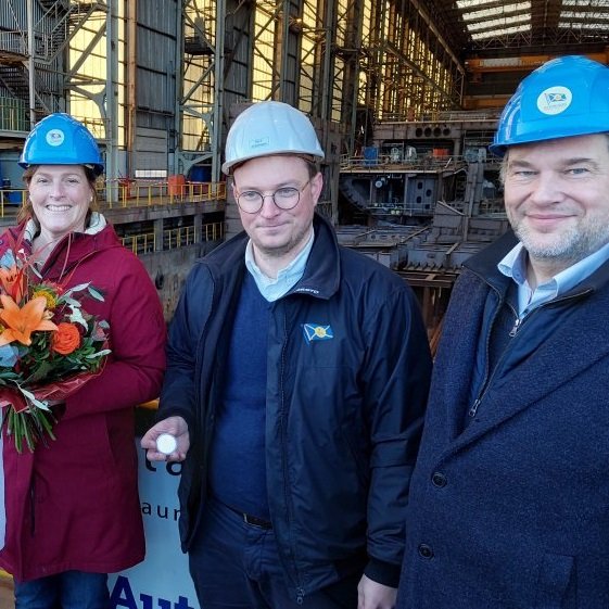 Zwei Männer und eine Frau, alle mittleren Alters, lächeln in die Kamera. Alle drei tragen blaue Bauhelme, die Frau hält einen bunten Blumenstrauß in den Händen.