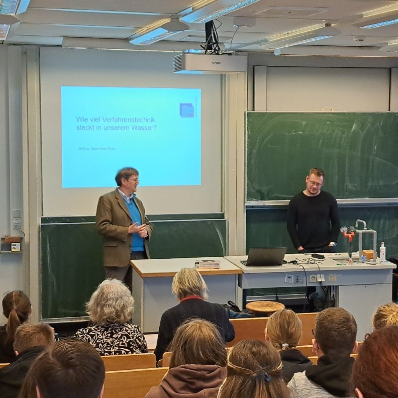 Ein junger und ein älterer Mann stehen vor einem Vorlesungssaal voller Menschen, hinter ihnen an der Wand eine Tafel und eine Projektion einer Präsentations-Titelfolie: "Wie viel Verfahrenstechnik steckt in unserem Wasser?"