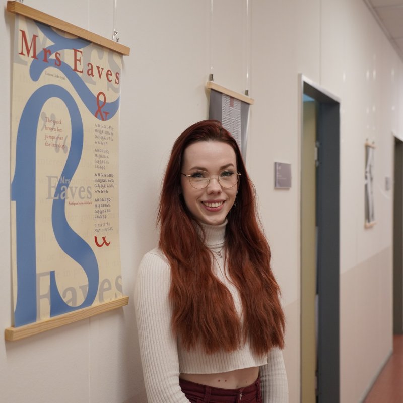 Eine junge Frau mit roten Haaren steht in einem Flur neben einem Plakat, auf dem ein großes ß abgebildet ist sowie der Schriftzug &quot;Mrs Eaves&quot; in verschiedenen Farben und weitere Schriftzüge. Im Hintergrund sieht man Türen und weitere Plakate.