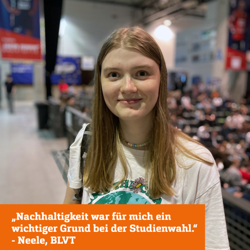 Eine junge Frau lächelt in die Kamera, sie trägt ein weißes T-Shirt mit dem Bild der Erdkugel, der grüne Schriftzug darüber wird halb von ihren Haaren verdeckt. Im Hintergrund sieht man verschwommen eine Halle und einen Rang, in dem viele weitere Menschen sitzen.
