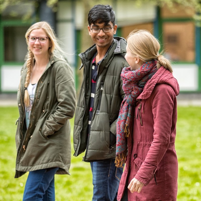 Zwei blonde junge Frauen und ein junger Mann mit dunklen Haaren und dunkler Haut laufen sich freundlich unterhaltend über den Flensburger Campus.