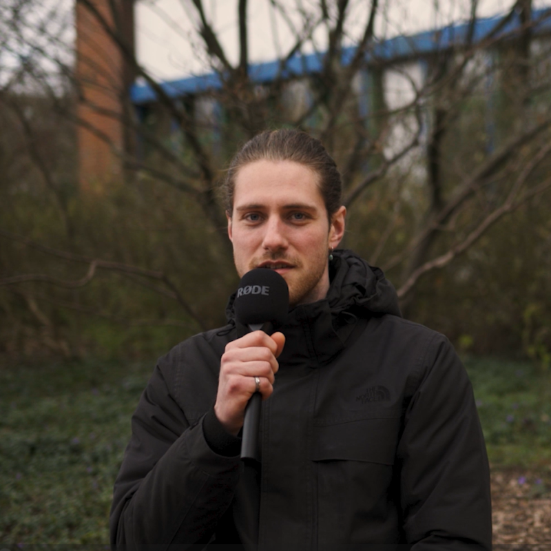 Ein juner Mann mit langen, zum Dutt gebundenen Haaren in einer dunklen Jacke spricht in ein Mikrofon, im Hintergrund sieht man einen Busch und eine Gebäudefassade.