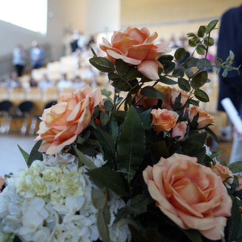 Im Vordergrund ein Blumenstrauß in weiß und rosa, im Hintergrund der Vorlesungssaal des Flensburger audimax, das sich gerade mit Menschen füllt.