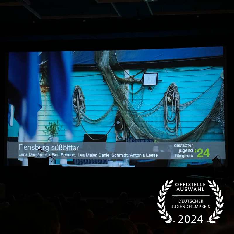 In einem schwarzen Kinosaal sieht man an der Leinwand ein Still aus einem Film mit einer blauen Holzwand, an der Taue hängen. Darunter steht der Text "Flensburg Süßbitter - Lena Dandanelle, Ben Schaub, Lea Majer, Daniel Schmidt und Antonia Leese".