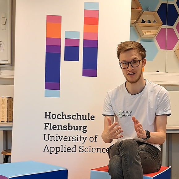 Ein junger Mann im weißen T-Shirt sitzt auf einem bunten Sitzwürfel und gestikuliert beim Sprechen. Hinter ihm steht ein Roll-Up mit dem Logo der Hochschule Flensburg.