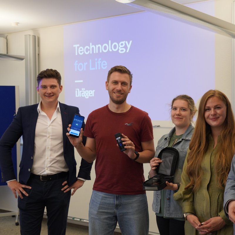 Drei Studierende und zwei Männer lächeln in die Kamera, im Hintergrund eine Projektion mit der Überschrift "Technology for Life - Dräger". Einer der Studierendne hält ein Smartphone und ein Messgerät, eine andere ein Schutzcase. 