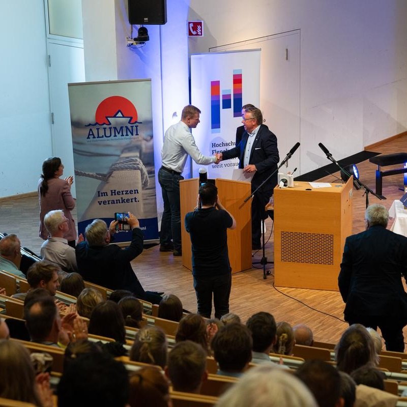 In einem voll besetzten Hörsaal steht ein Mann im Anzug an einem Rednerpult und schüttelt einem jüngeren Mann im blauen Hemd die Hand. Hinter ihnen stehen zwei Roll-Ups mit den Logos der Hochschule Flensburg und des Alumni-Vereins.