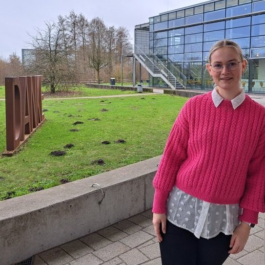 Eine junge Frau in einem leuchtend rosafarbenen Pullover, unter dem eine weiße Bluse hervorschaut, lehnt an einer Säule. Im Hintergrund sieht man ein verglastes Gebäude, Bäume und eine grüne Wiese, auf der in Großbuchstaben aus rostbraunem Metall das Wort DANKE steht.