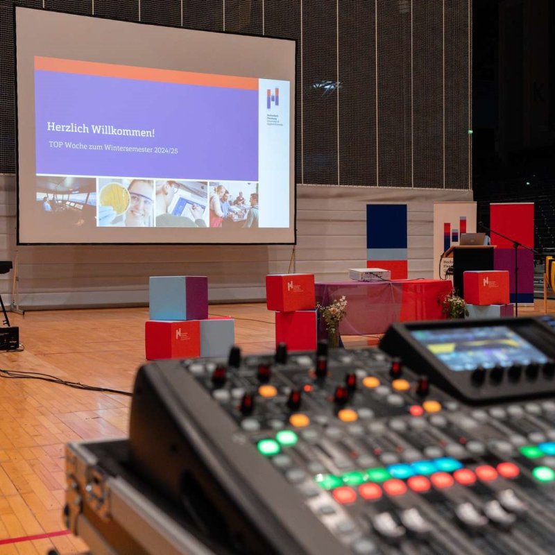 Nahaufnahme eines Tonmischpults, im Hintergrund eine Bühne mit vielen bunten Würfeln, Rollups und einer Leinwand mit der Projektion: Herzlich Willkommen! TOP Woche zum Wintersemester 2024/25"
