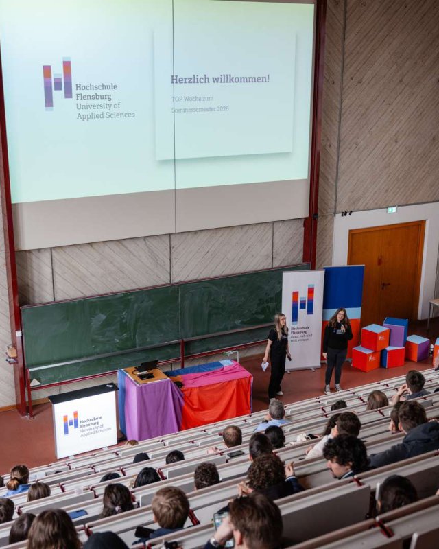 In einem gut gefüllten Vorlesungssaal stehen vorne zwei Studentinnen mit Mikrofonen, hinter ihnen Text an der Wand: Herzlich willkommen zum Sommersemester 2026.