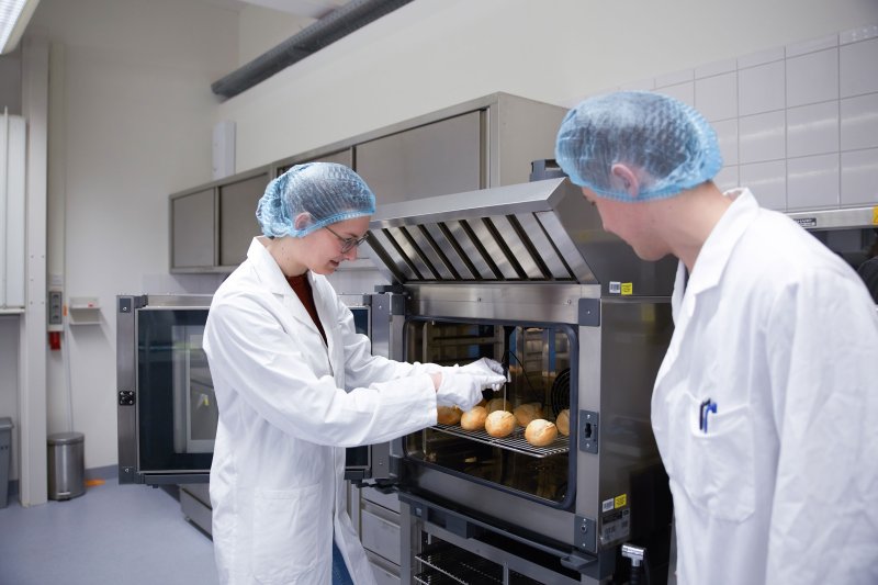 Zwei Studierende in Laborkittel und mit Haarschutz vor einem Ofen, sie führen eine Messung durch.
