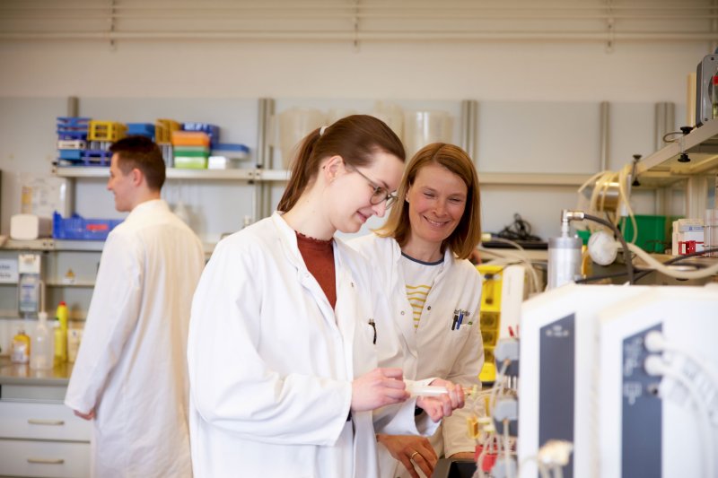 Eine Studentin und eine Dozentin im Labor, sie stehen vor einer Anlage, beide tragen Laborkittel.
