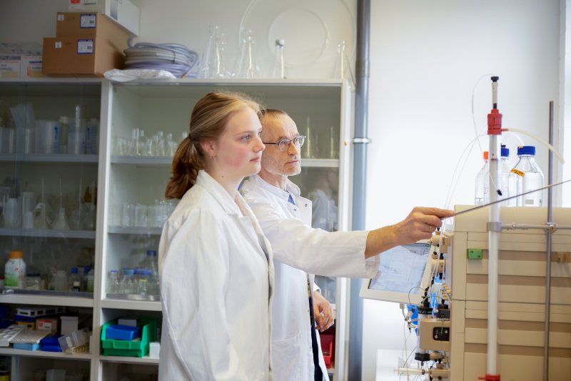 Eine Studentin und ein Dozent im Labor, beide tragen Laborkittel. Er zeigt mit einem Zeigestock auf etwas.