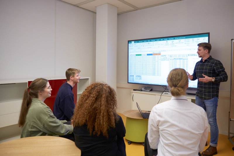 Seminarszene: Studierende sitzen vor einem großen Bildschirm. Ein Student erklärt etwas.