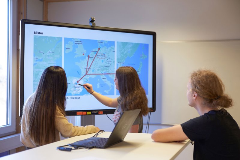 Drei Studierende vor einem Bildschirm mit einer Weltkarte. Eine Studentin zeigt etwas.