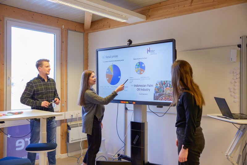 Drei Studierende stehen um einen Bildschirm, eine Studentin zeigt etwas. Auf dem Bildschirm sind Diagramme zu erkennen.