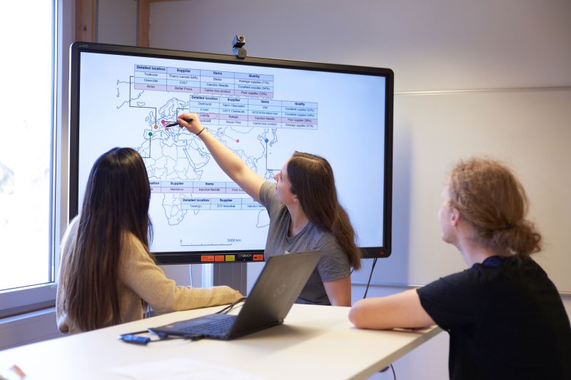 Drei Studierende vor einem großen Bildschirm. Zu erkennen ist eine Karte und Tabellen, eine Studentin zeigt etwas.