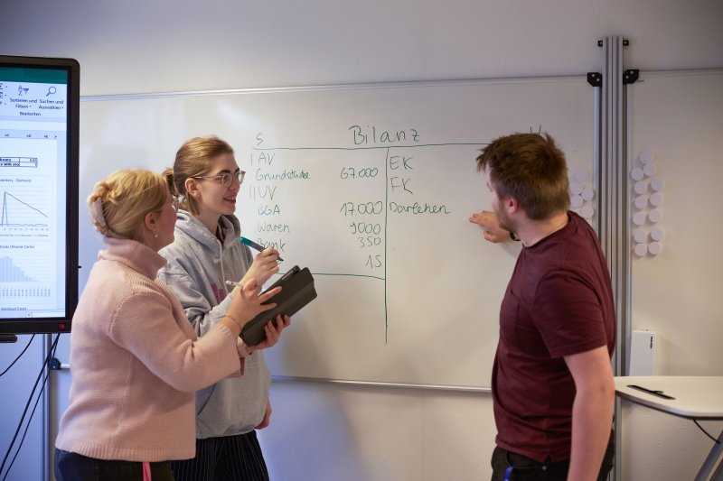 Drei Studierende vor einem Whiteboard, einer zeigt darauf, die anderen machen sich Notizen. Auf dem Boad ist das Wort "Bilanz" zu erkennen.
