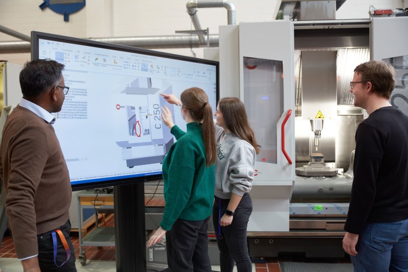 Studierende un Dozent vor einer Spritztechnik-Fertigungsanlage. Zwei Studentinnen zeigen auf ein 3D-Model an einem Bildschirm.