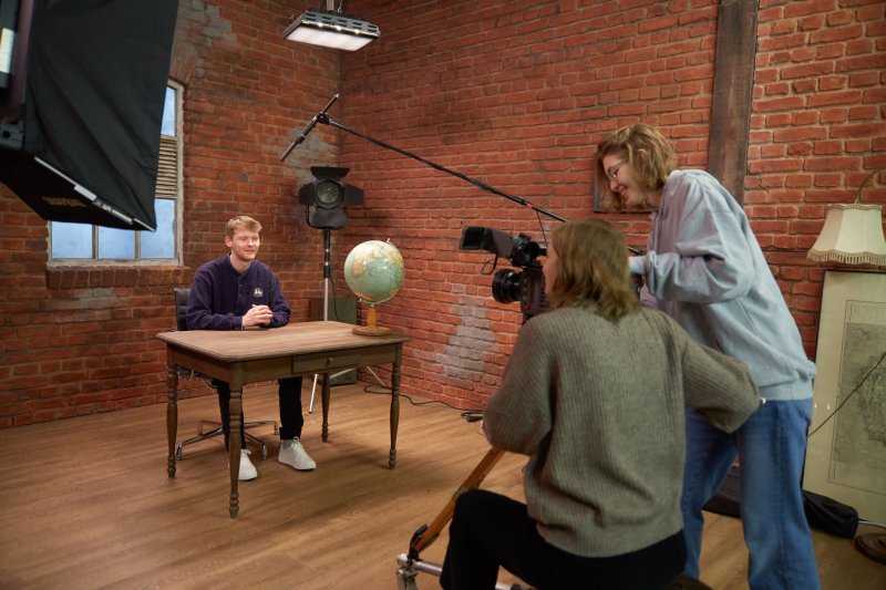 TV Studio: Ein Student sitzt an einem Tisch, er wird von zwei Studentinnen an der kamera gefilmt.