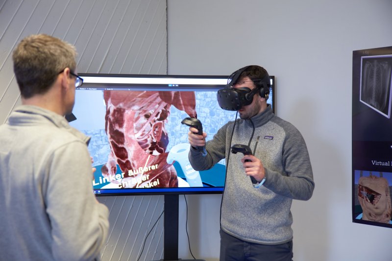 Zwei Menschen vor einem Bildschirm, einer trägt eine VR Brille, auf dem Bilschirm ist ein Oberkörper, Sehenen, Rippen etc. zu erkennen.