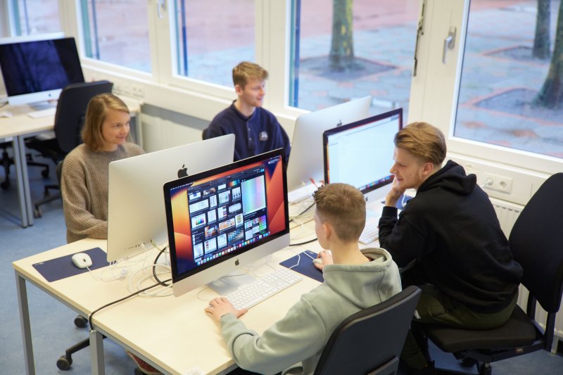 Vier junge Menschen an einer Arbeitsinsel mit Rechnern und Bildschirmen.