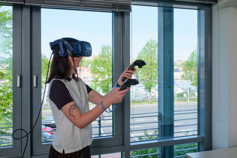 Eine Studentin trägt eine VR Brille und hält in der Hand einen Controller. Hinter ihr eine große Fensterfront mit Blick ins Grüne.