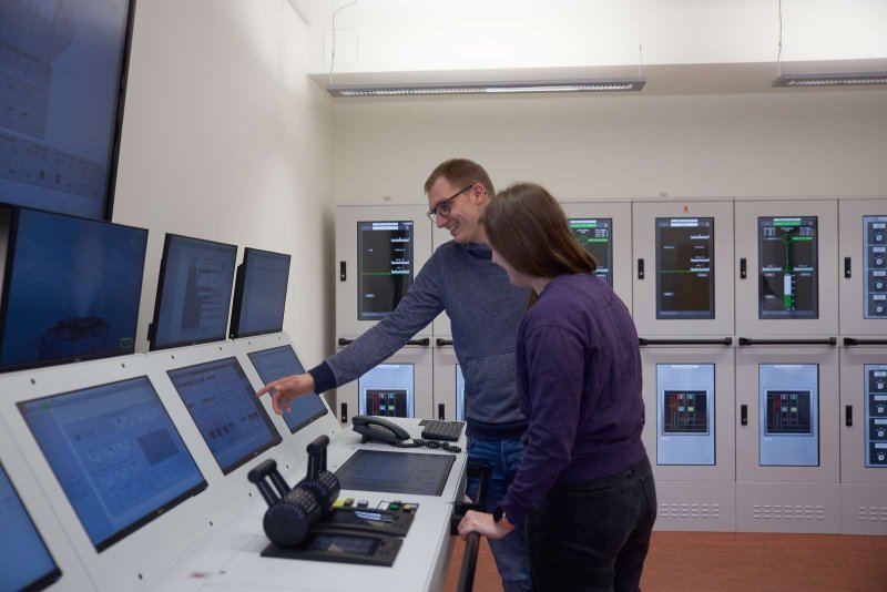 Eine Studentin und ein Student stehen an einem Schiffsmaschinen-Simulator, einem Panel mit eingelassenen Bildschirmen, Hebeln und einem Telefon. Der Student zeigt auf etwas auf einem Monitor und beide lächeln.