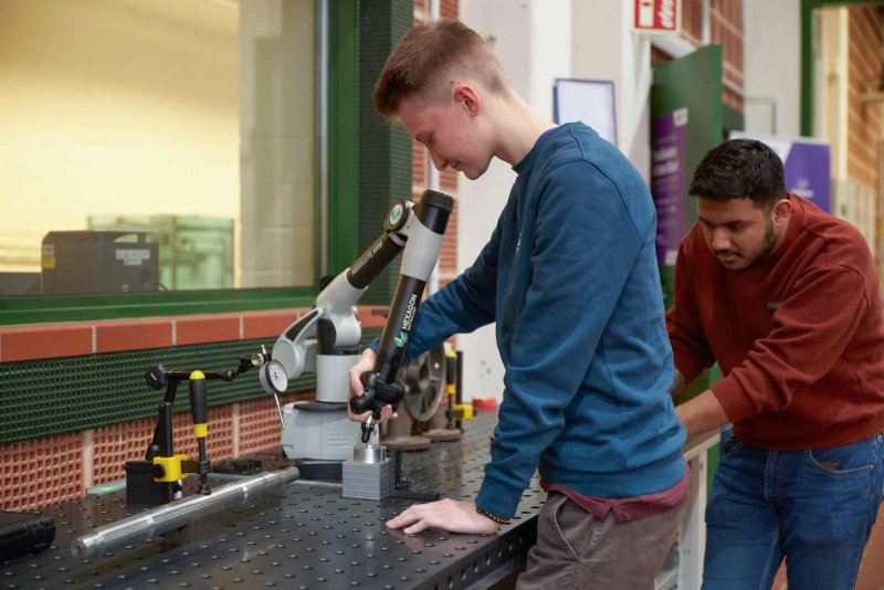 Zwei Studenten stehen an einem Metallpult und bearbeiten Metallteile. Einer blickt durch ein Mikroskop auf das Metallteil, der andere beugt sich vor, um so mehr zu sehen.