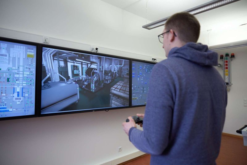 Ein Student steht vor einer Reihe von an der Wand aufgehängten Monitoren. Rechts und links sieht man die schematische Darstellung eines Maschinenraums, auf dem mittleren Monitor die virtuelle Nachbildung des Maschinenraums. Der Student hält einen Controller in der Hand und steuert durch den virtuellen Raum.