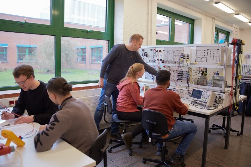 Vier Studierende stehen in einem Elektrotechnik-Labor an einer elektrischen Schaltvorrichtung und lassen sich etwas von einem Dozenten erklären.