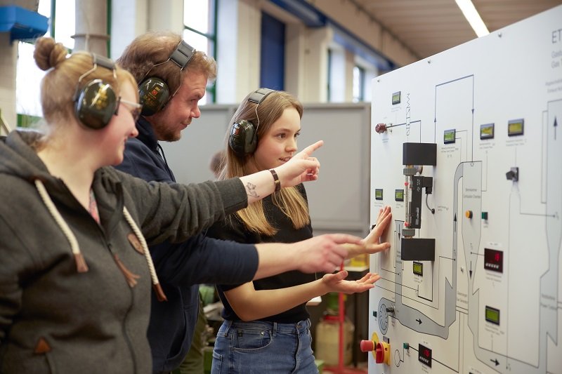 Drei Studierende stehen vor einer elektrischen Schaltvorrichtung, eine dreht an einem Knopf und eine andere zeigt auf etwas.