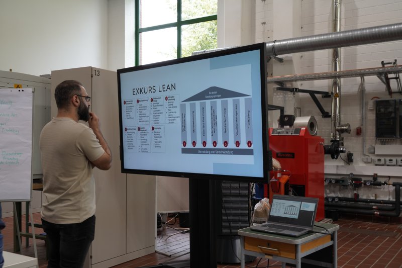 Ein Studierender steht vor einem Monitor, auf dem ein Diagramm mit der Übrschrift "Exkurs Lean" zu sehen ist. Im Hintergrund mehrere technische Gerätschaften und Rohre.