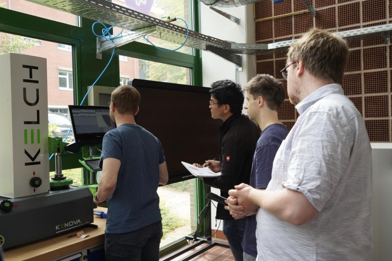 In einer Maschinenhalle mit großer Fensterfront stehen vier Studierende vor einem großen Monitor, einer von ihnen tippt gerade etwas an einem Rechner daneben ein, die anderen blicken ihm konzentiert über die Schulter.