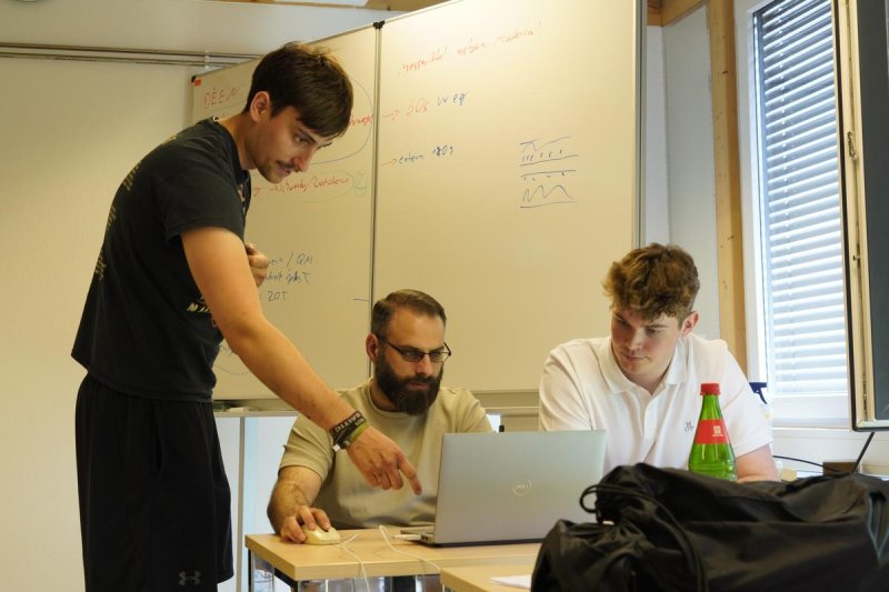 Zwei Studenten sitzen in einem Seminarraum und blicken konzentriert auf einen Laptop-Schirm, ein dritter steht hinter ihnen und zeigt auf den Laptop.